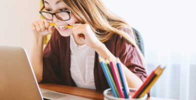 woman biting pencil while sitting on chair in front of computer during daytime